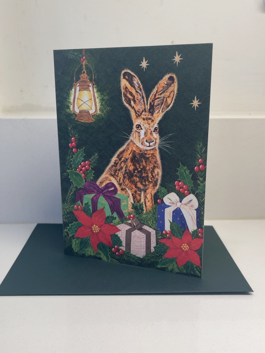 Christmas Wishes art card featuring a hare, glowing lantern and presents, with its dark green envelope