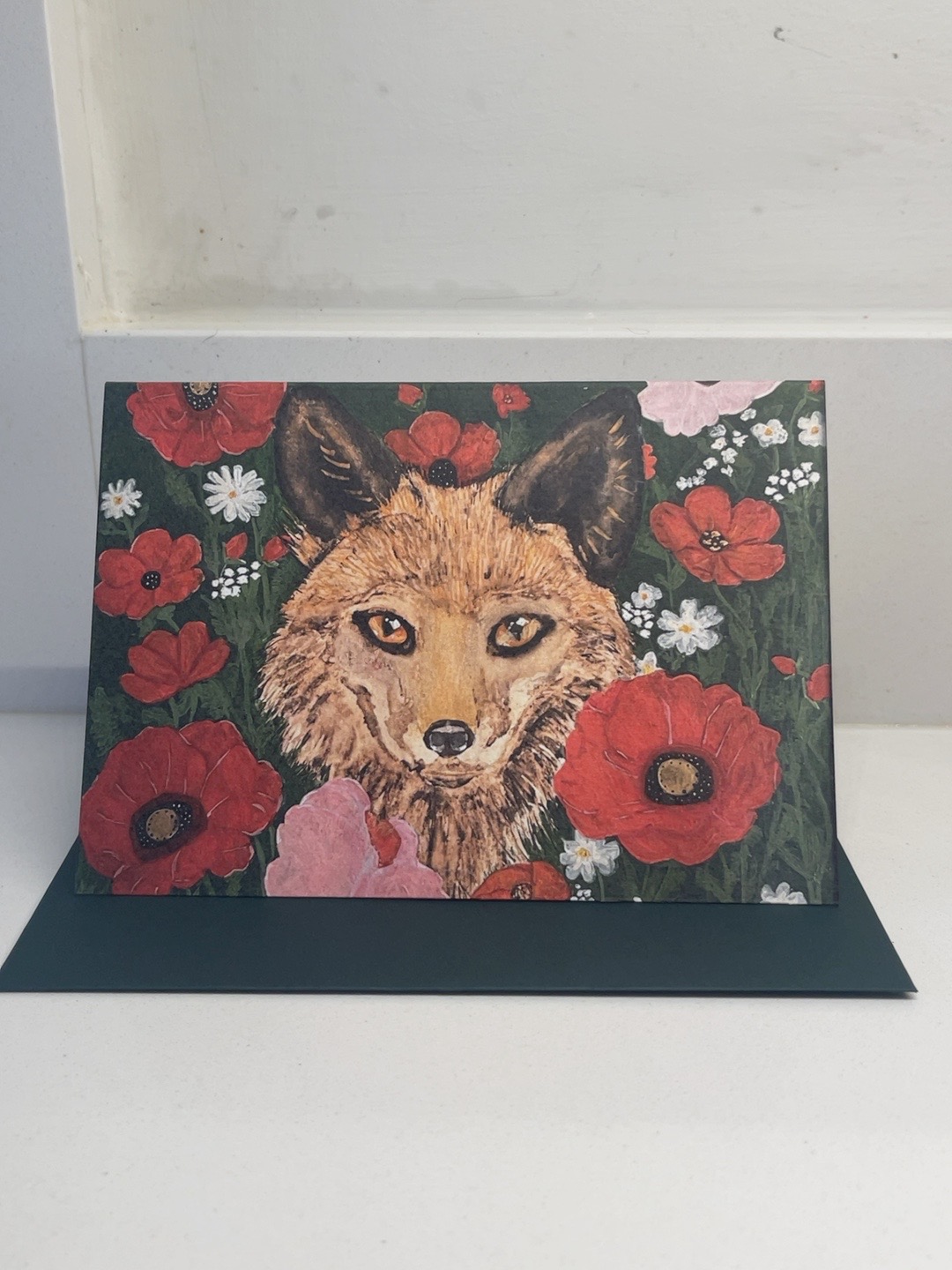 Rose-Tinted Shelter fox and flowers art card, photographed with its dark green envelope